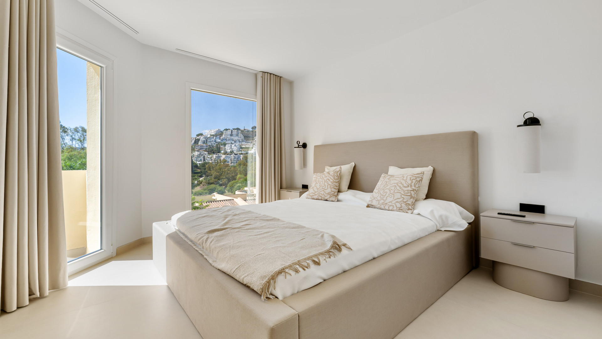 Bright bedroom with two windows and mountain views. Beige bed and curtains enhance the light ambiance.