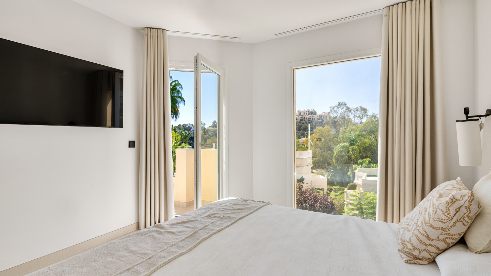 Cozy bedroom featuring a wall-mounted TV and views of trees from two open windows. Light curtains add softness.