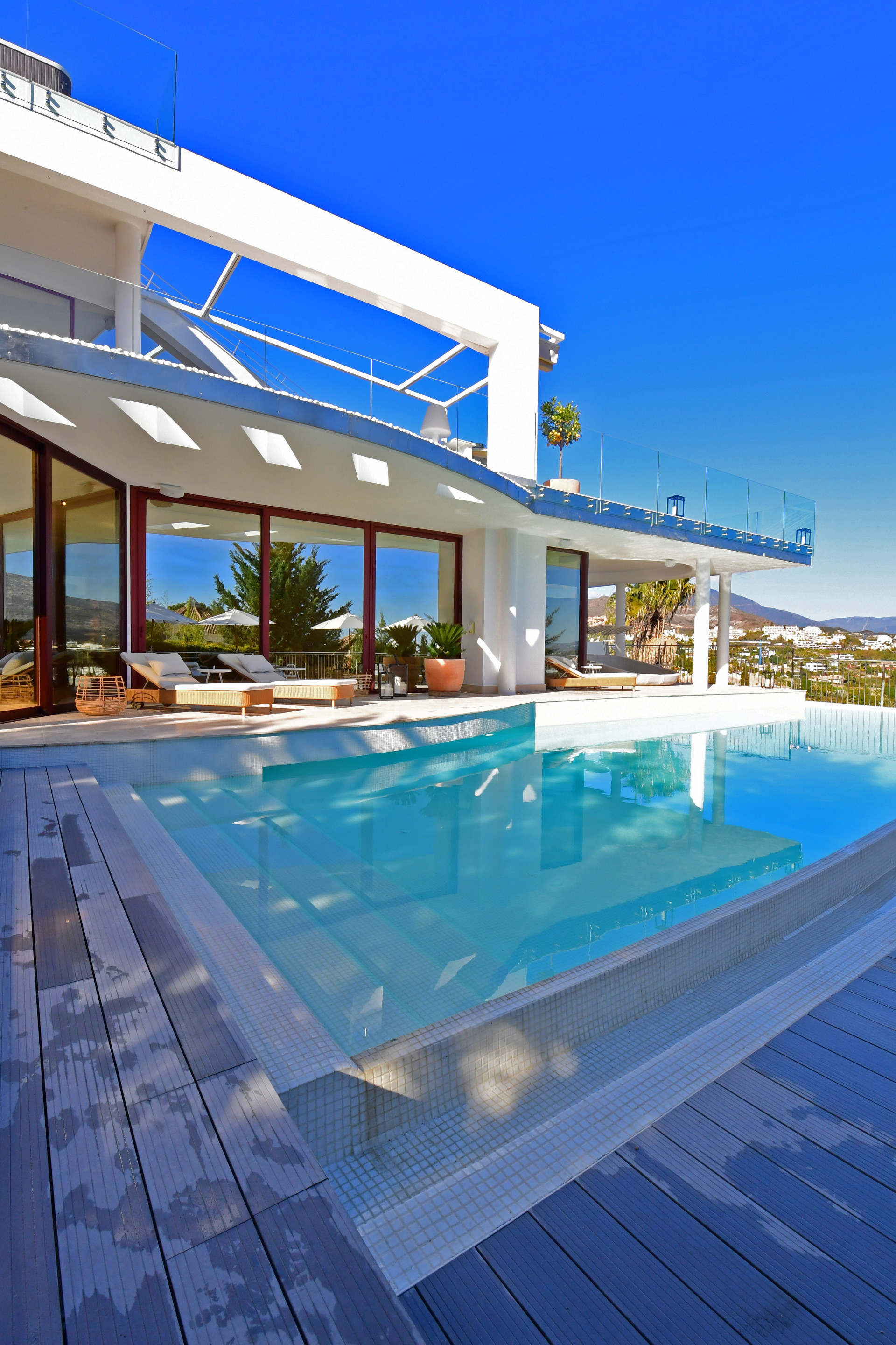 View of the pool area with sun loungers and large windows showcasing the house interior.