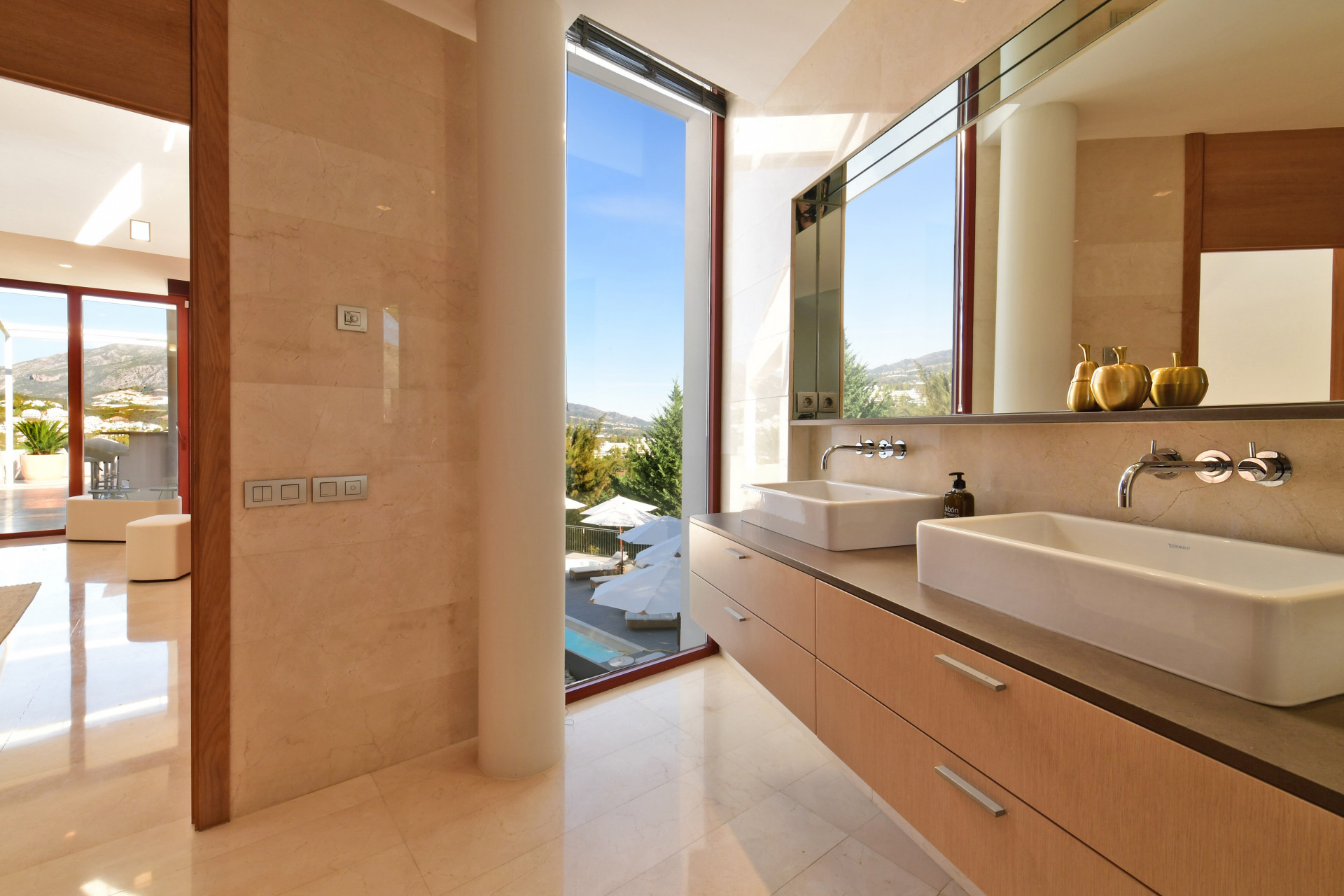 Elegant bathroom with dual sinks, marble surfaces, glass accents, and a view of the outdoor area.