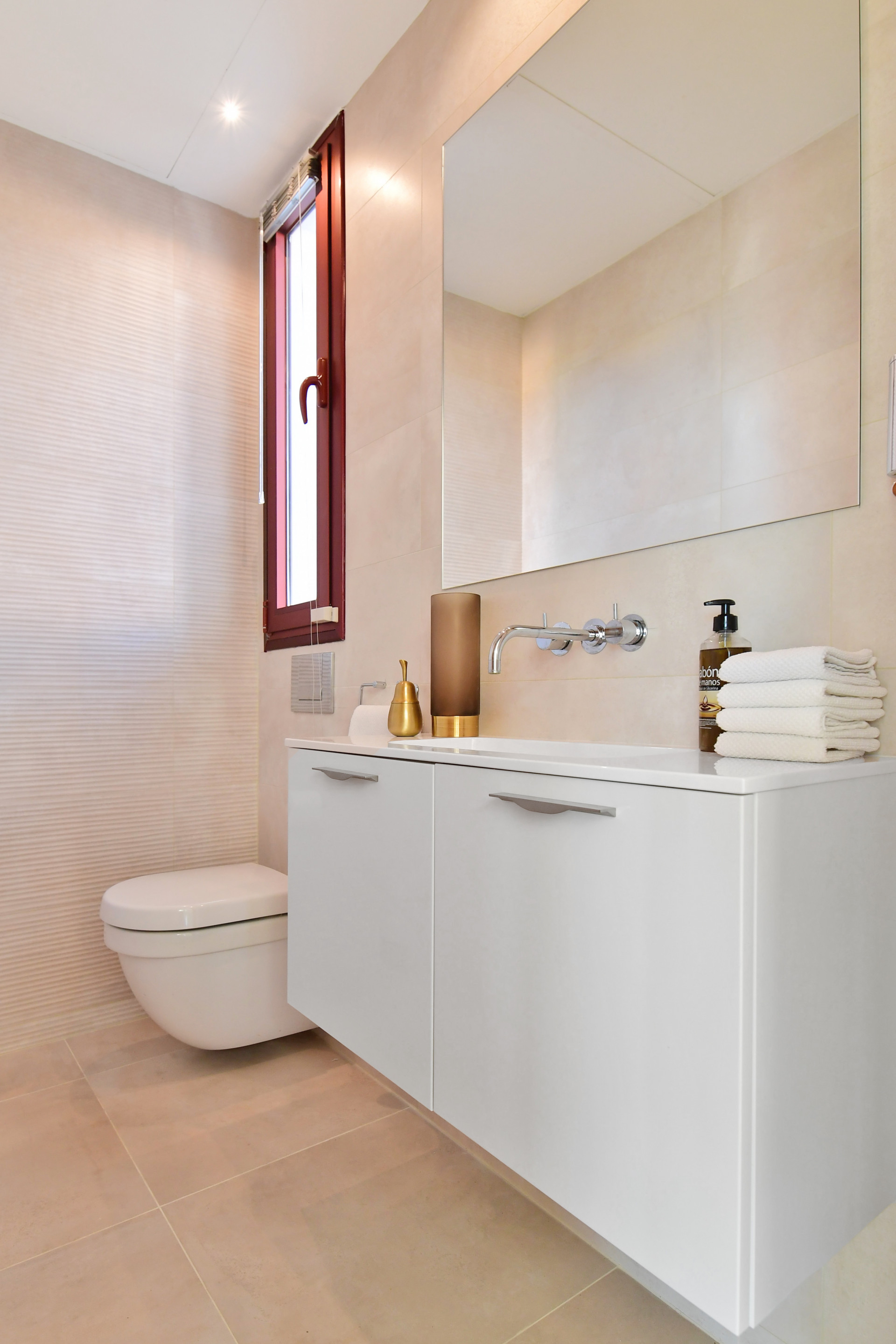 Modern bathroom with beige tones, white vanity, and large mirror beside a small window.