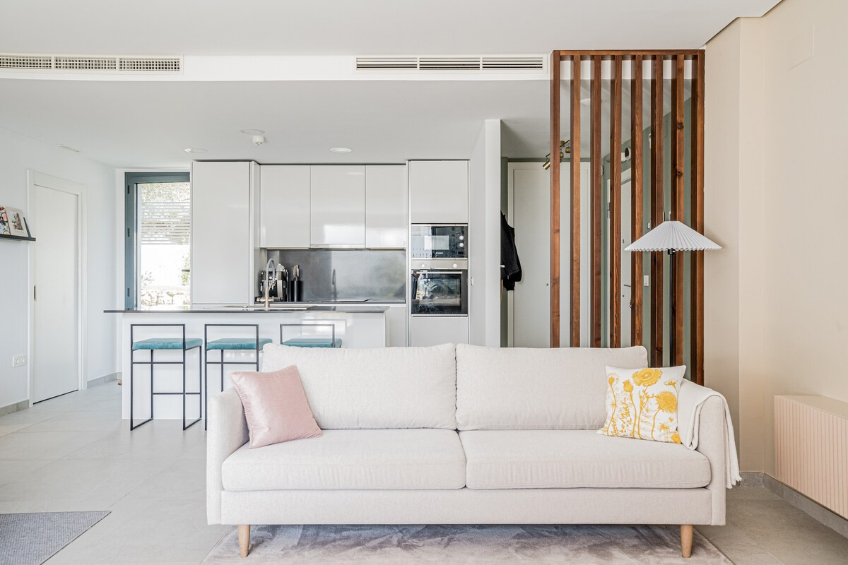 Open concept living space with a light sofa, wooden room divider, and modern kitchen visible in the background.