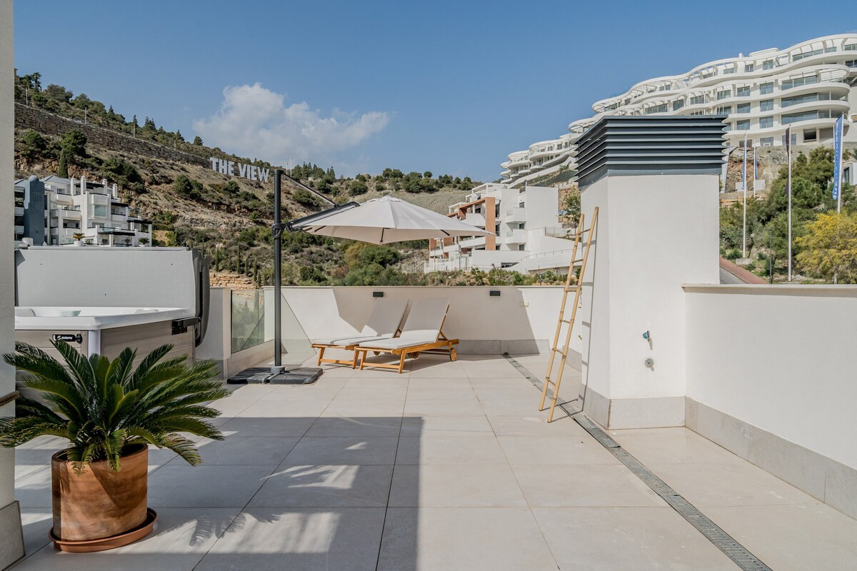 Rooftop terrace with two lounge chairs under an umbrella, hot tub, and mountain backdrop.