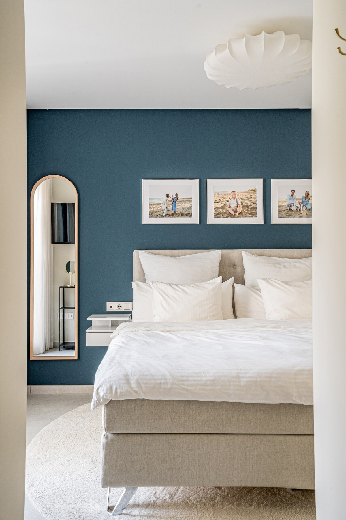 View from a doorway into a bedroom showing a bed, a full-length mirror, and blue wall with framed art.