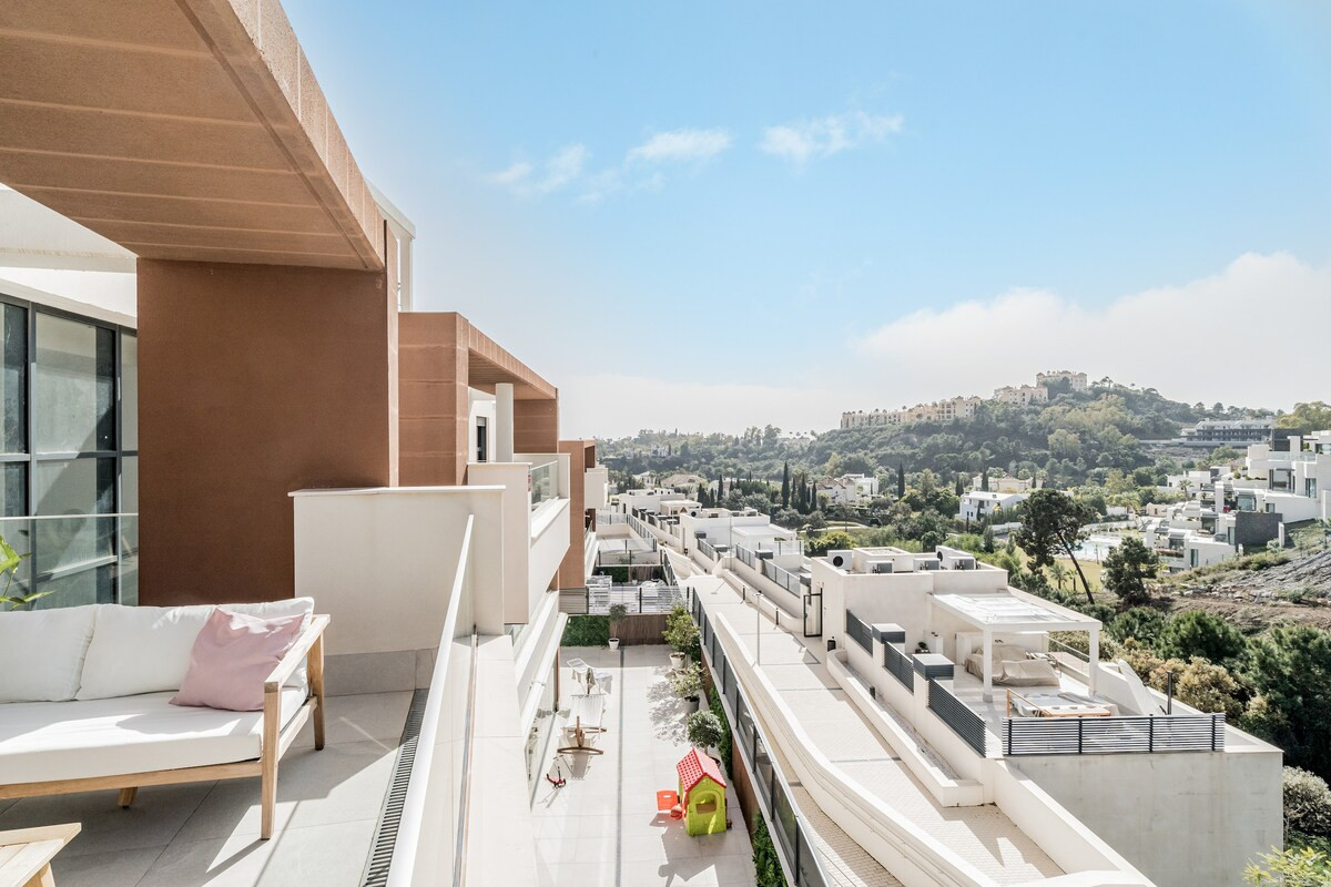 Outdoor balcony with white sofa and pink pillow overlooking a modern residential complex and distant hills.