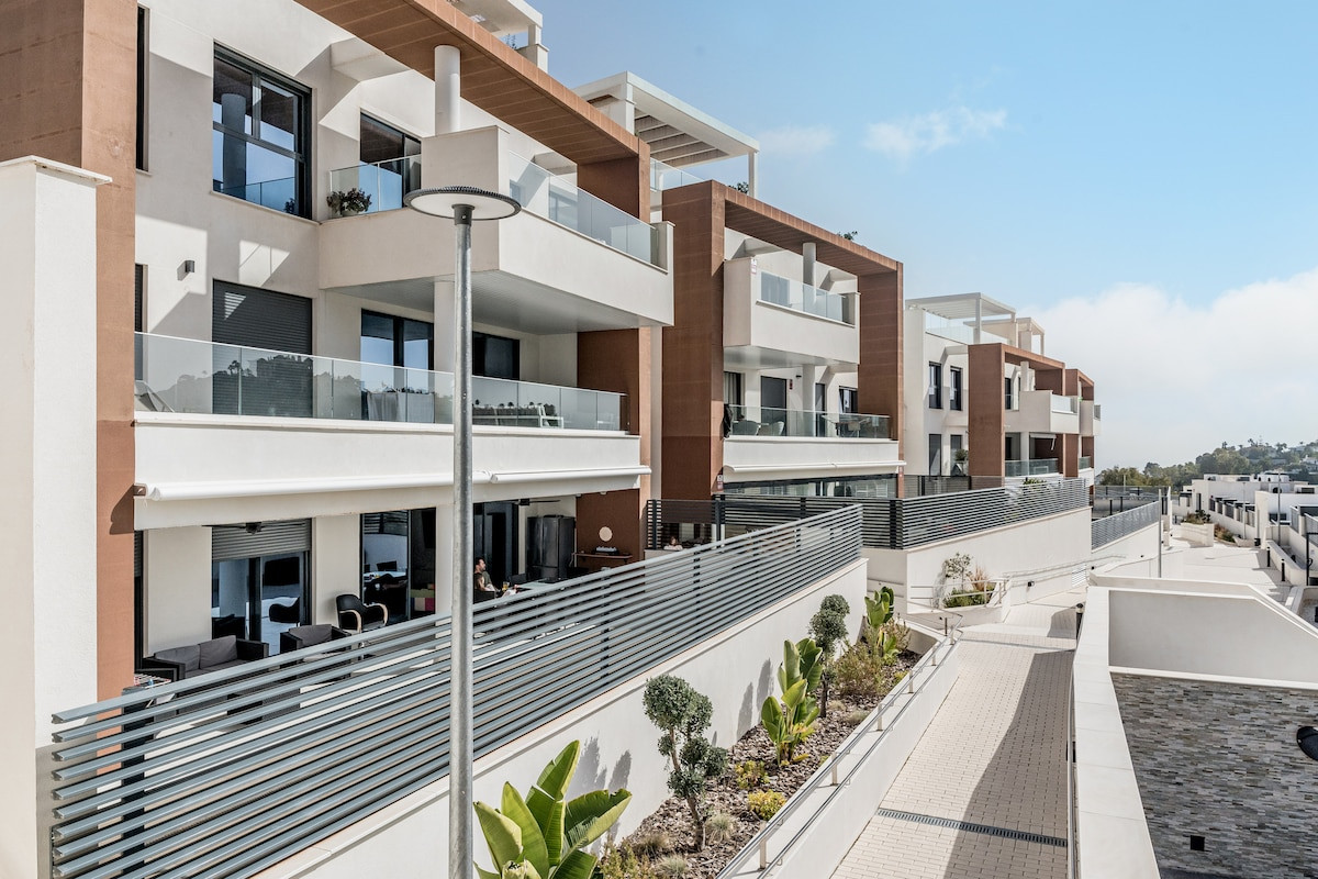 Sunny exterior of a multi-story apartment building with balconies and a paved pathway.