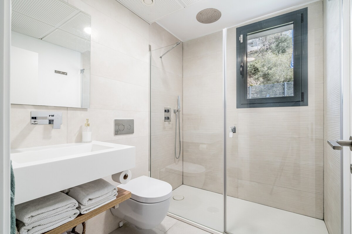 Bathroom with a floating vanity, toilet, glass shower, and window overlooking trees.