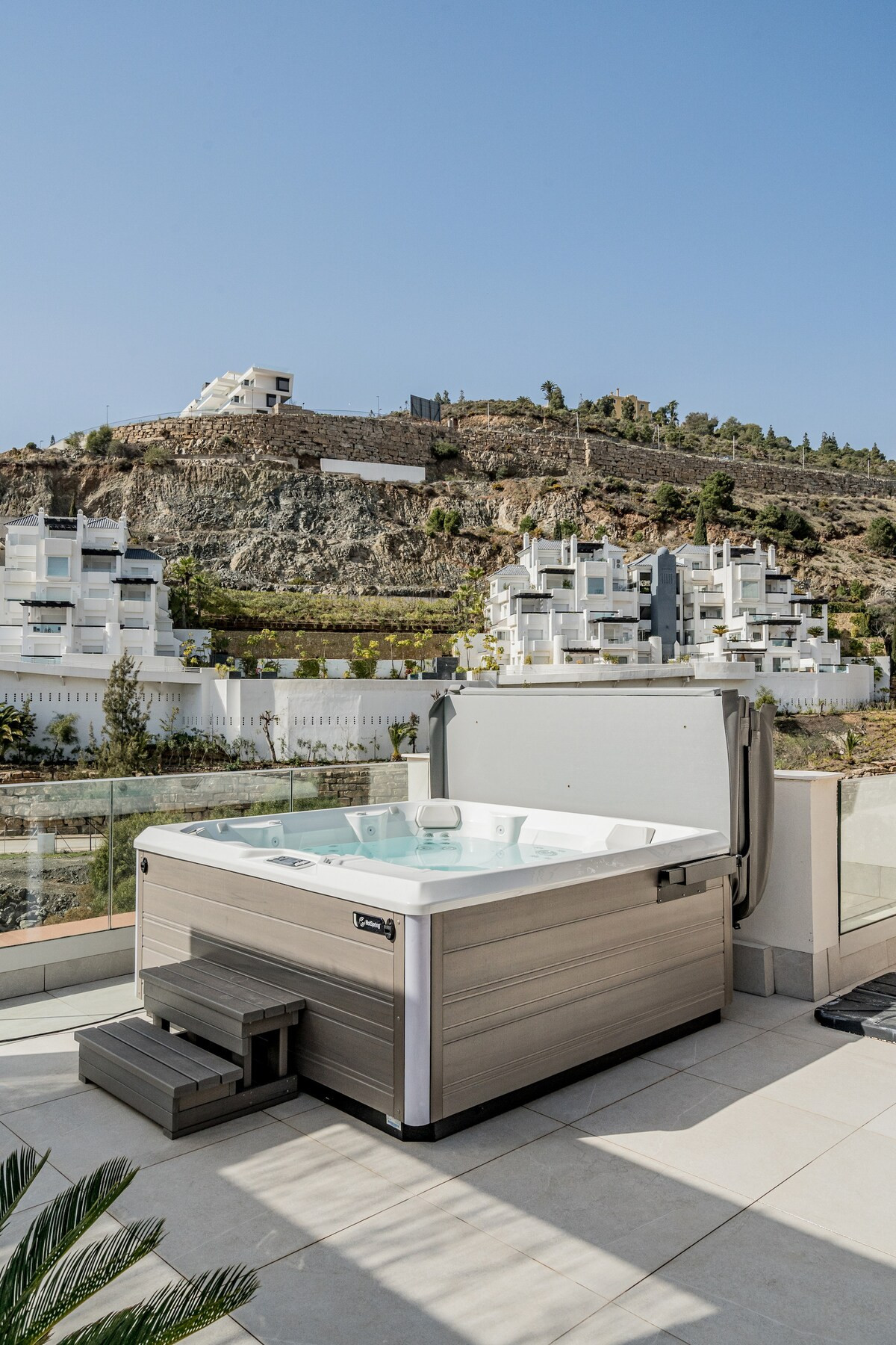 Outdoor hot tub on a tiled patio, with a hillside and buildings in the background.