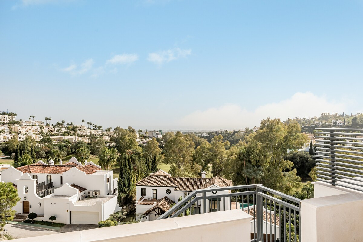 Expansive view from a balcony overlooking a residential area with traditional white houses and green trees.