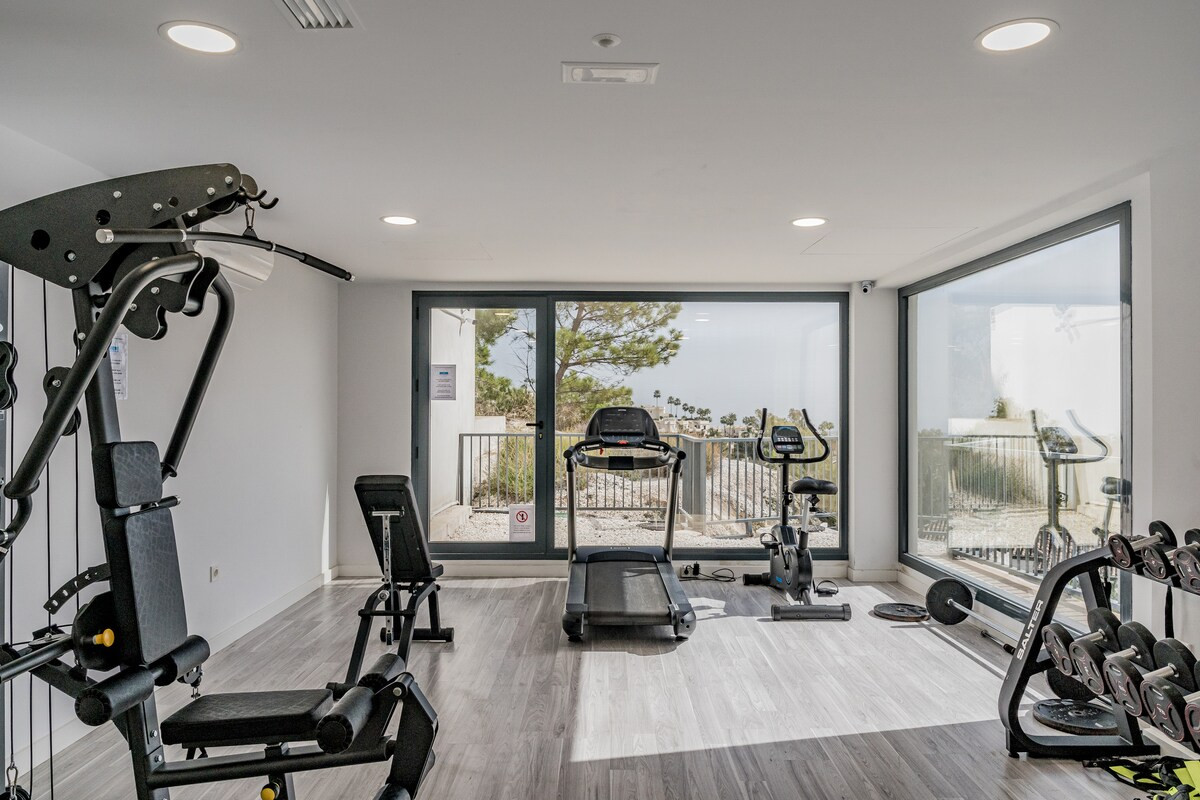 Indoor gym with a treadmill, exercise bike, weight bench, and a window overlooking an outdoor area.