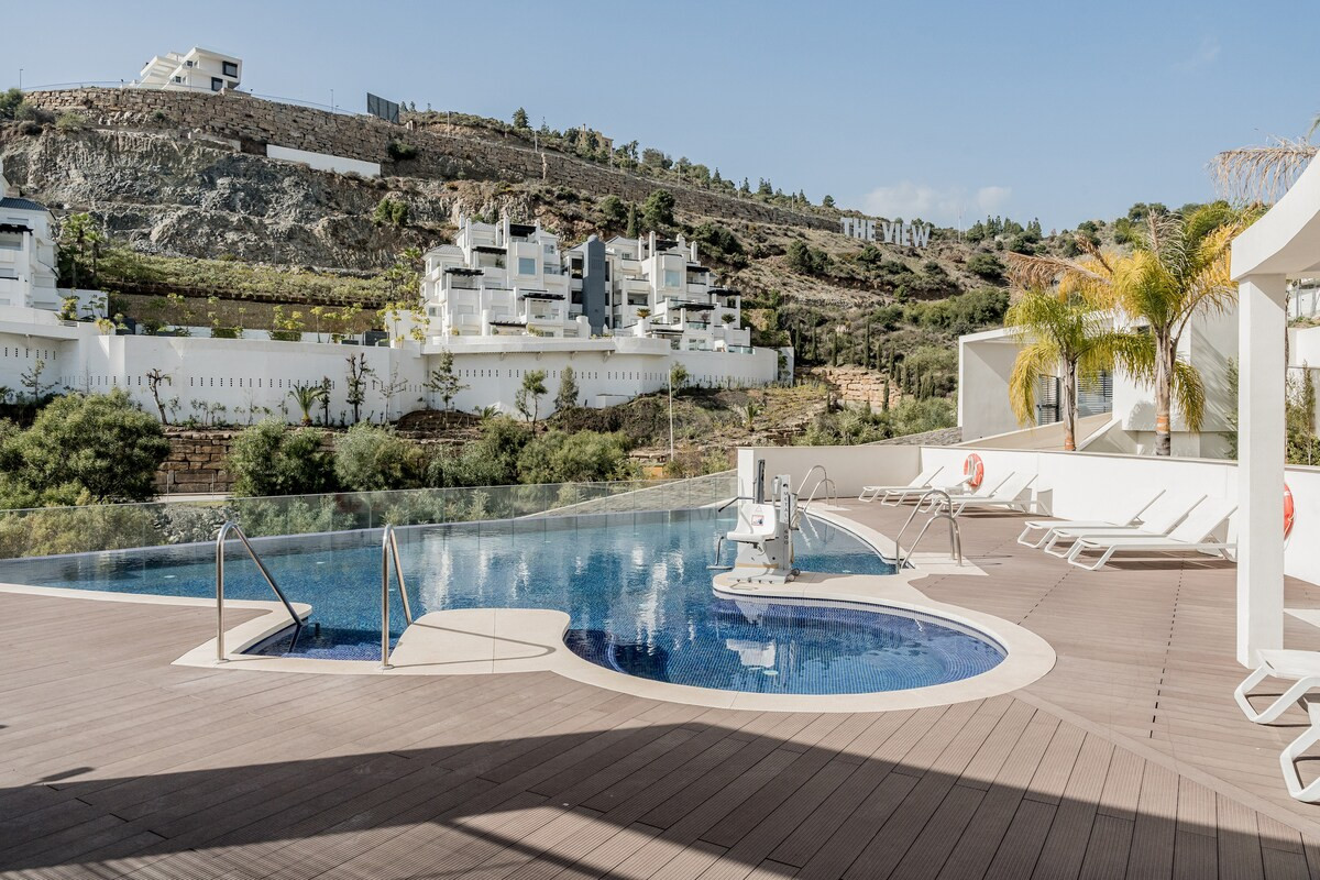 Large community swimming pool with a shallow circular section, surrounded by lounge chairs and a wooden deck.