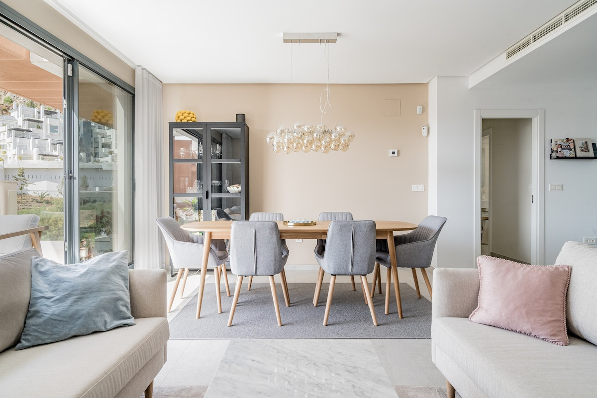 Dining area with a light wooden table, six grey chairs, a decorative chandelier, and a black display cabinet.