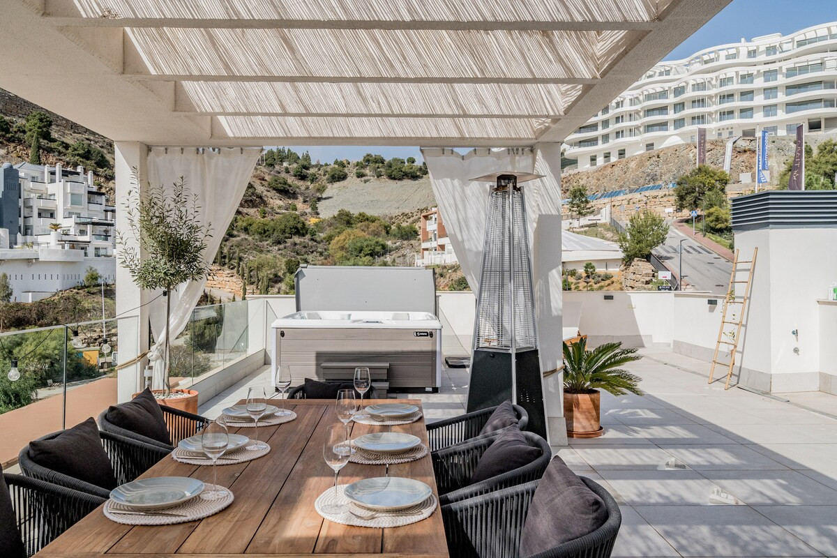 Outdoor terrace with a wooden dining table, a hot tub, and views of distant buildings and mountains.