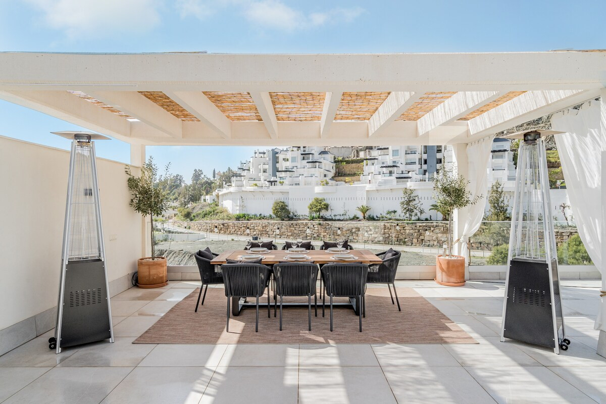 Outdoor dining area under a pergola with a large table and chairs, flanked by two heaters.