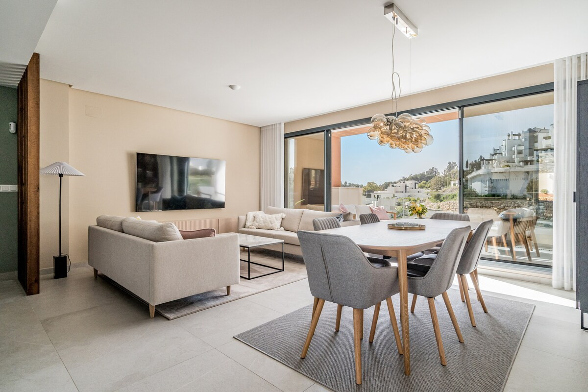 Spacious open living and dining area with a large wall-mounted TV, two light sofas, and glass doors to a terrace.