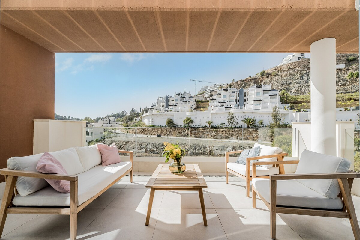 Outdoor lounge area on a patio with a sofa, chairs, and a table, overlooking a hillside.