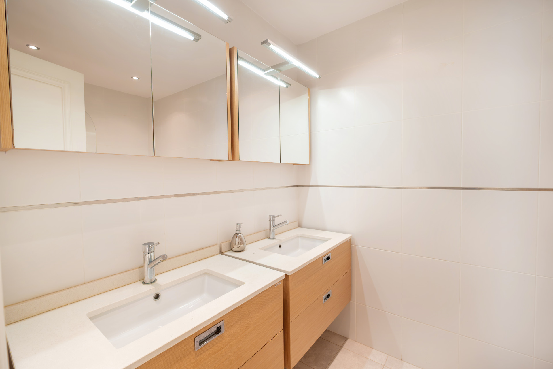 Modern bathroom with dual sinks and illuminated mirrors, featuring wooden cabinets.