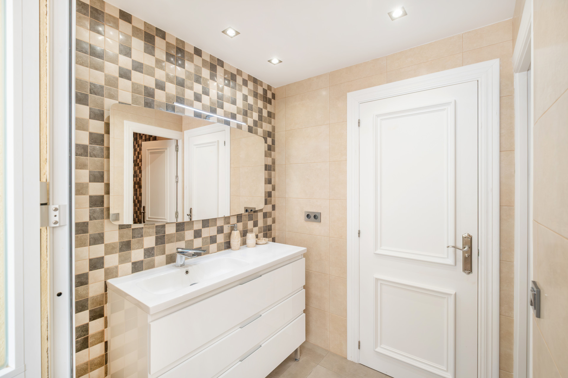 Modern bathroom vanity with a large mirror, checkered tiles, and an adjoining door leading to another room.
