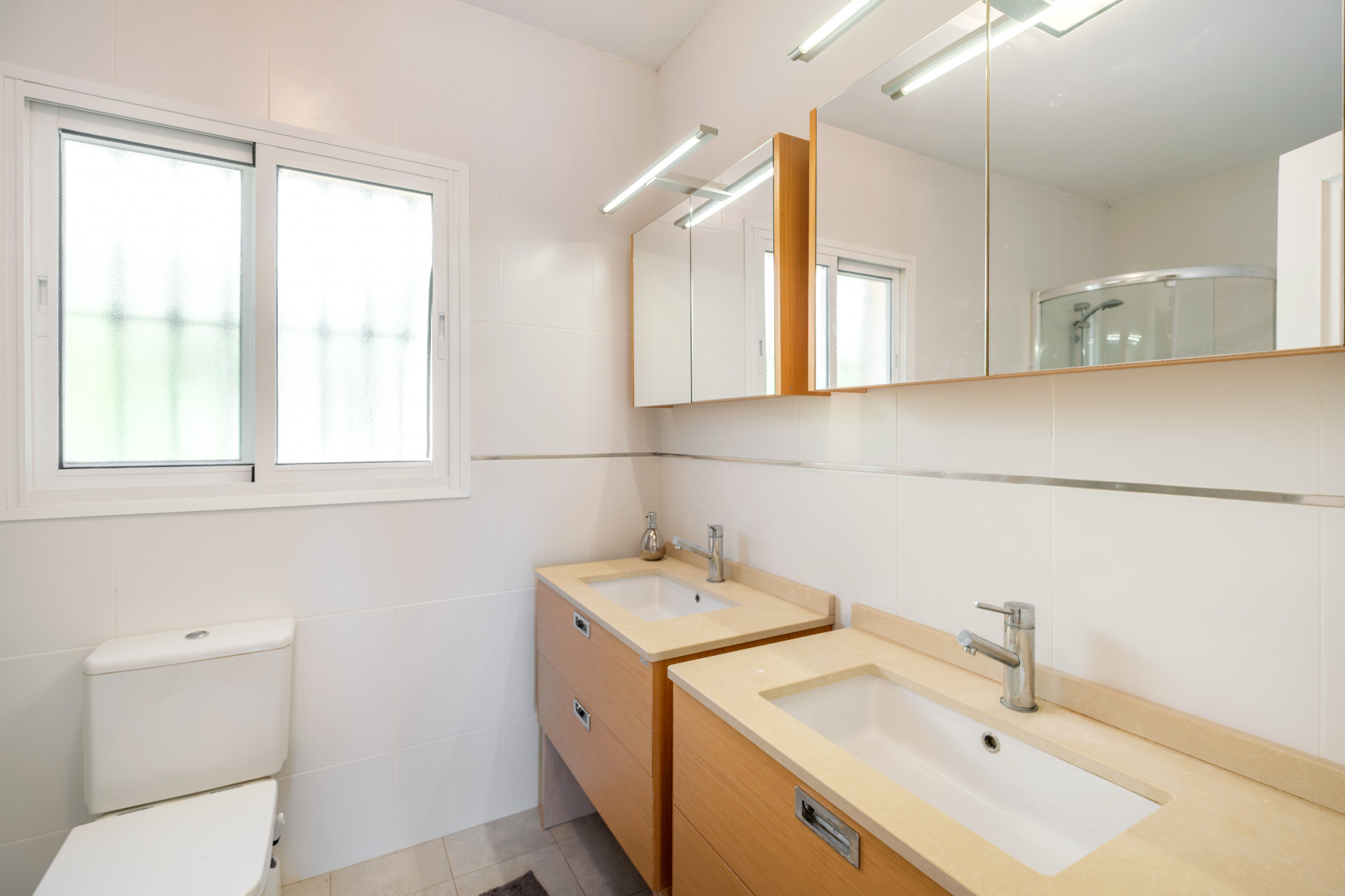 Well-lit bathroom including a dual sink, frosted window, and shower area.