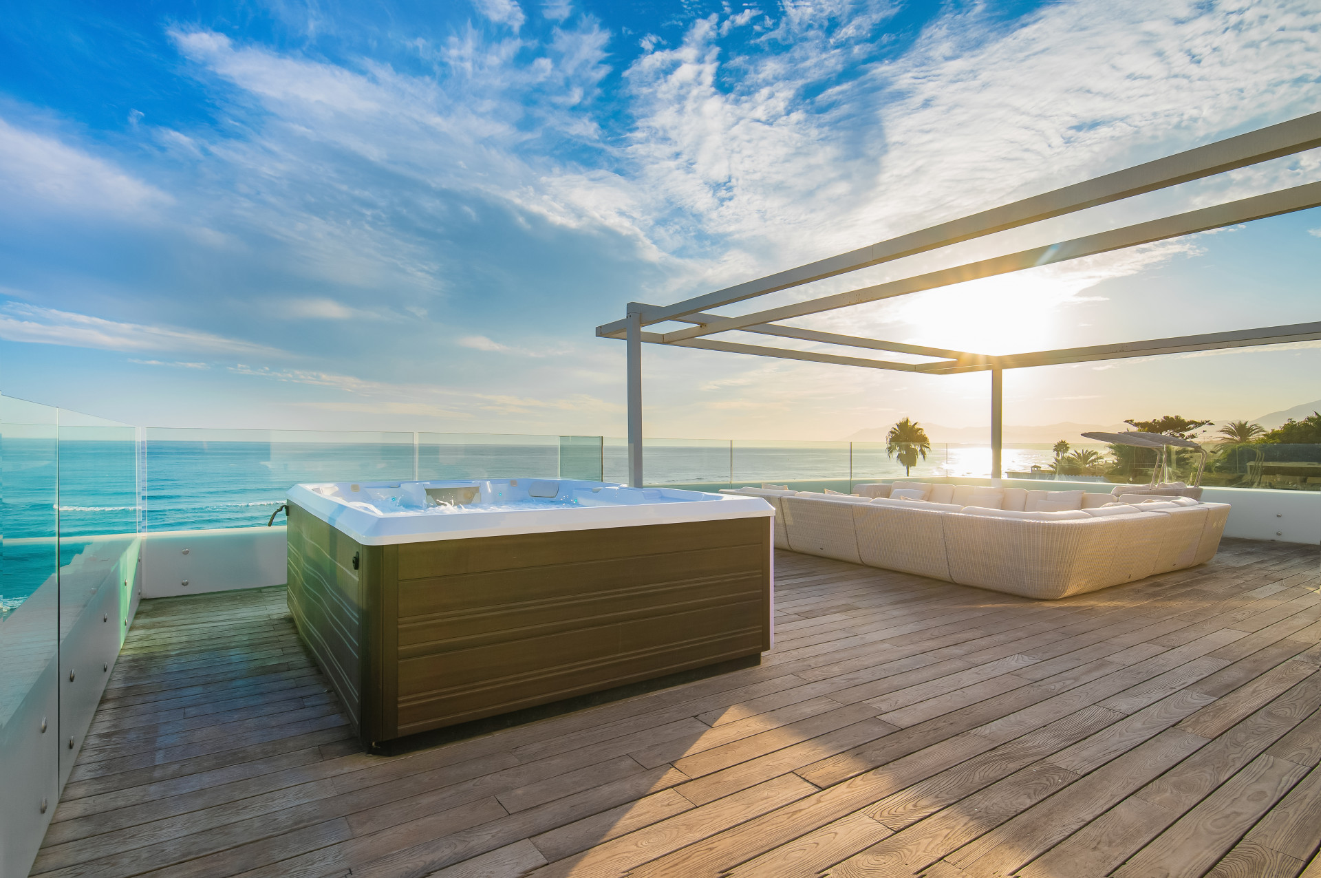 Jacuzzi on a wooden deck overlooking the ocean with a glass railing and a sunset sky.
