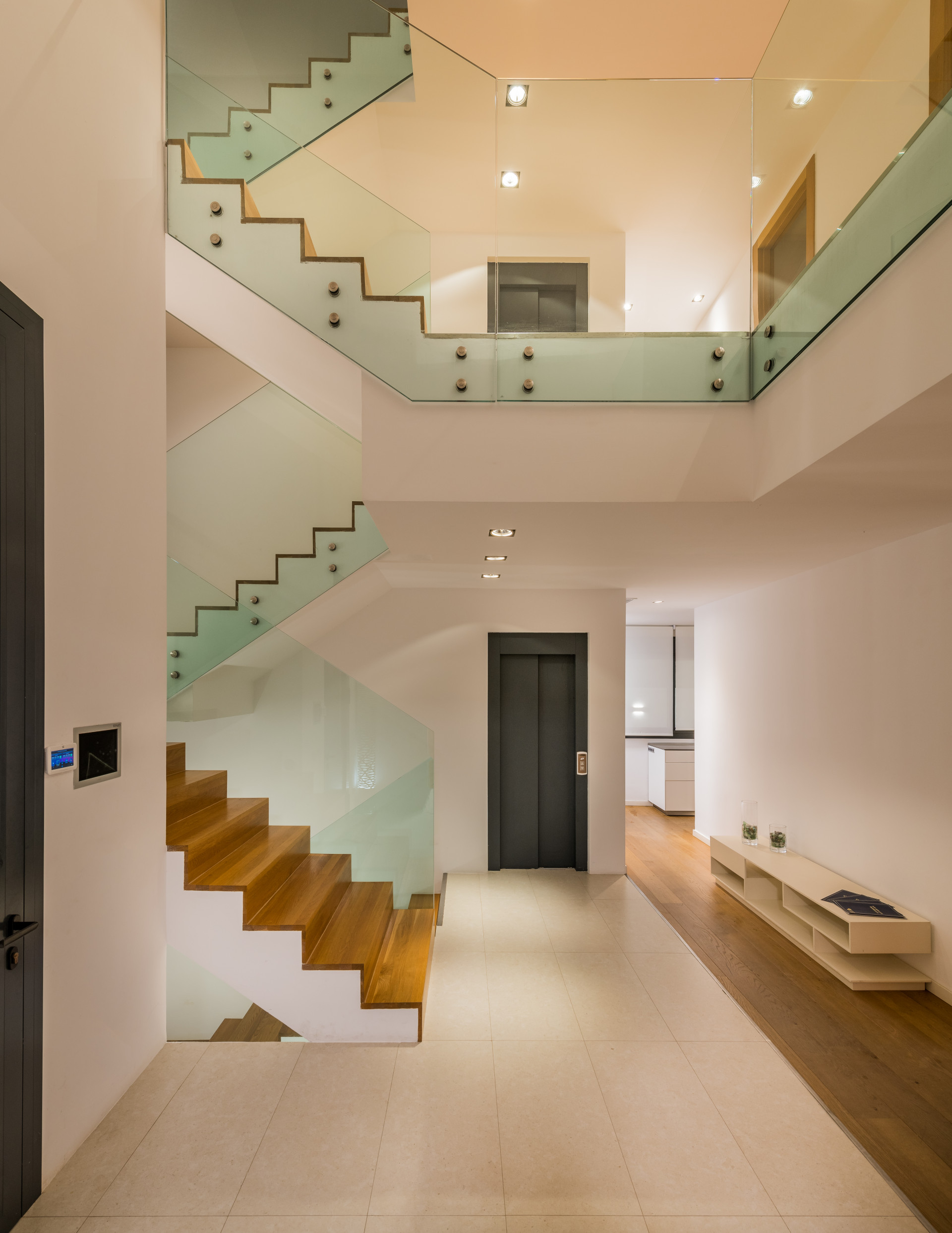 Modern staircase with glass railings and wooden steps, leading to an elevator and kitchen area.