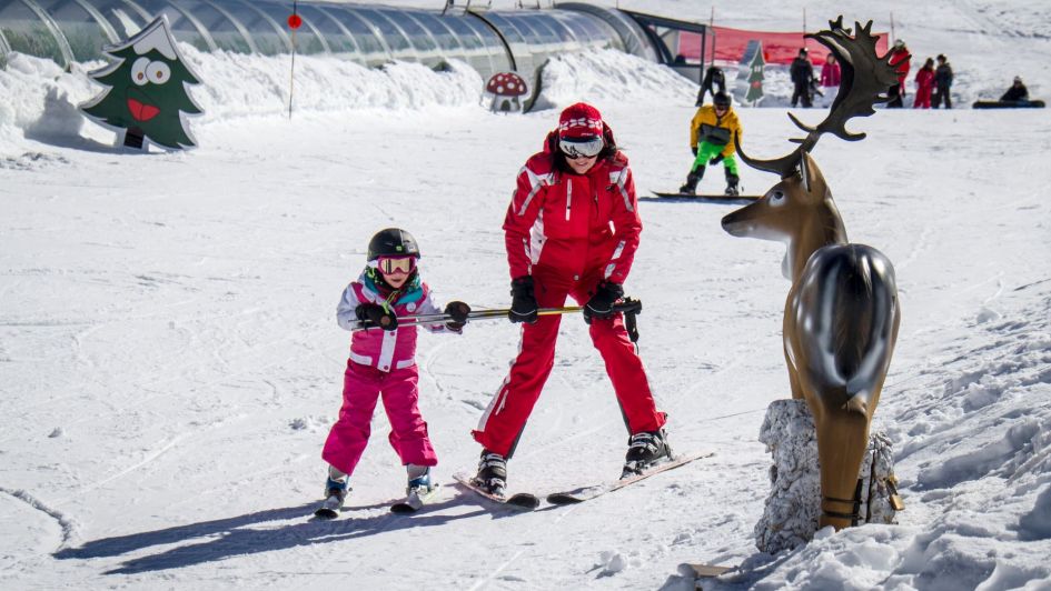 Little girl child having ski lesson in Sierra Nevada at Christmas time. showing the enclosed conveyor lift at the botton of the slopes, and a reindeer statue.